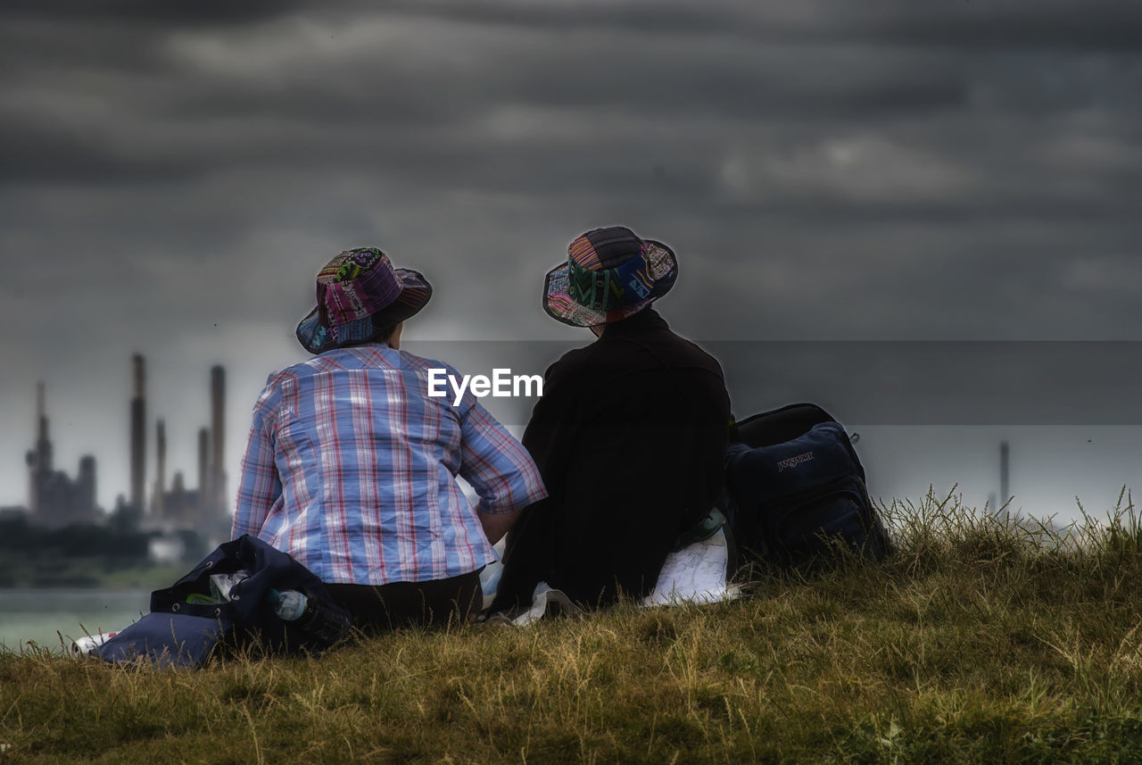 Rear view of couple sitting on grassy field against cloudy sky