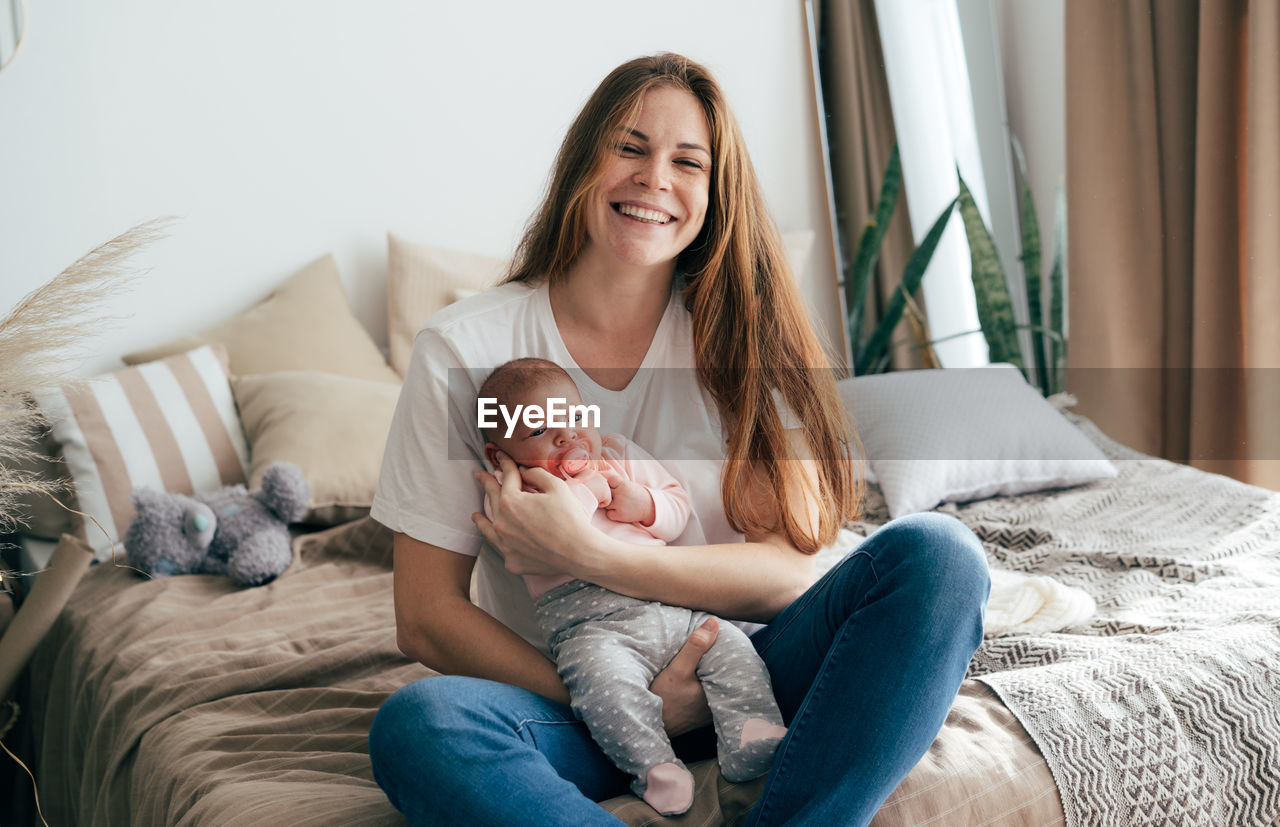 A young laughing mother holds a newborn baby in her arms while sitting on the bed.