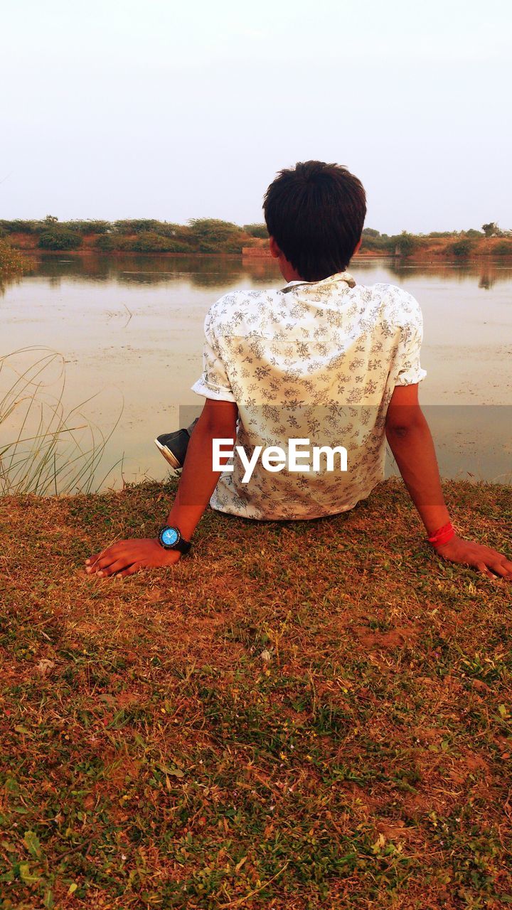 Rear view of man sitting on riverbank against clear sky at sunset