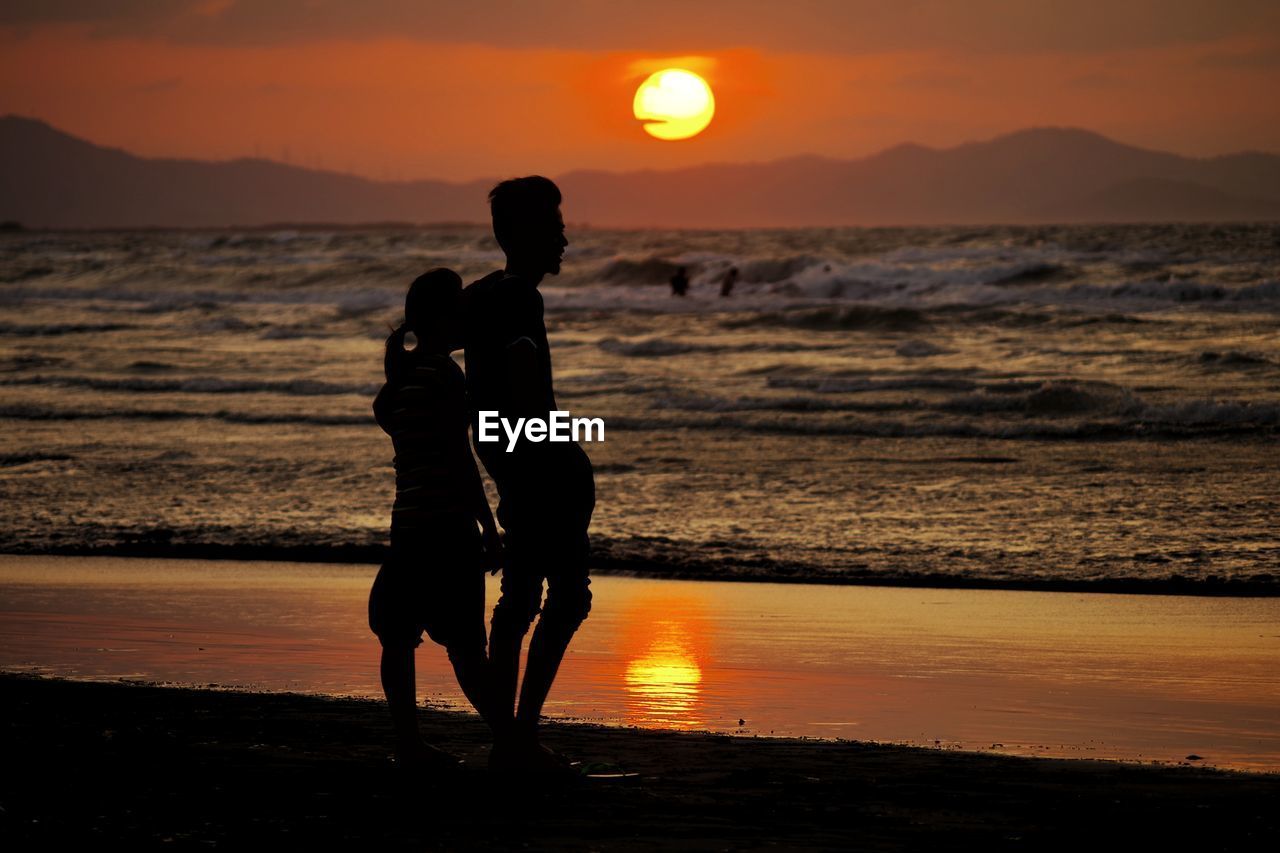 SILHOUETTE FRIENDS STANDING ON BEACH AGAINST ORANGE SKY