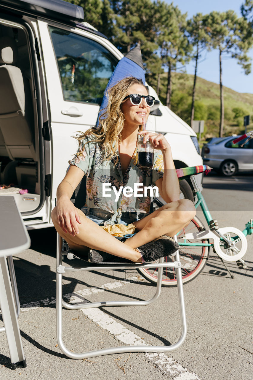 Cheerful woman enjoying a juice while sitting out of caravan in camping near surf board and bicycle