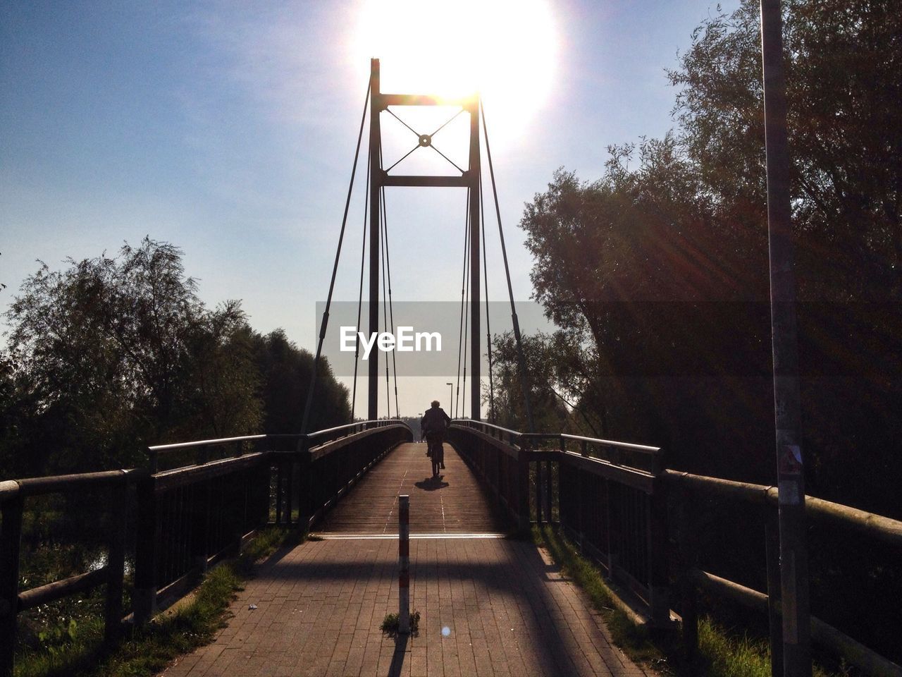 Silhouette person riding bicycle on bridge during sunny day