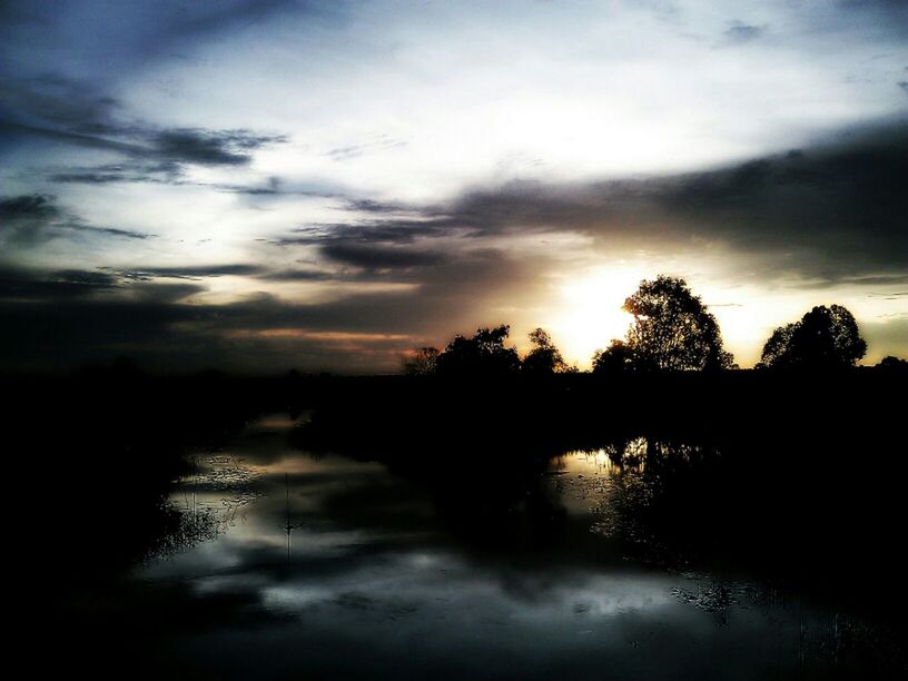 Lake by silhouette plants against cloudy sky