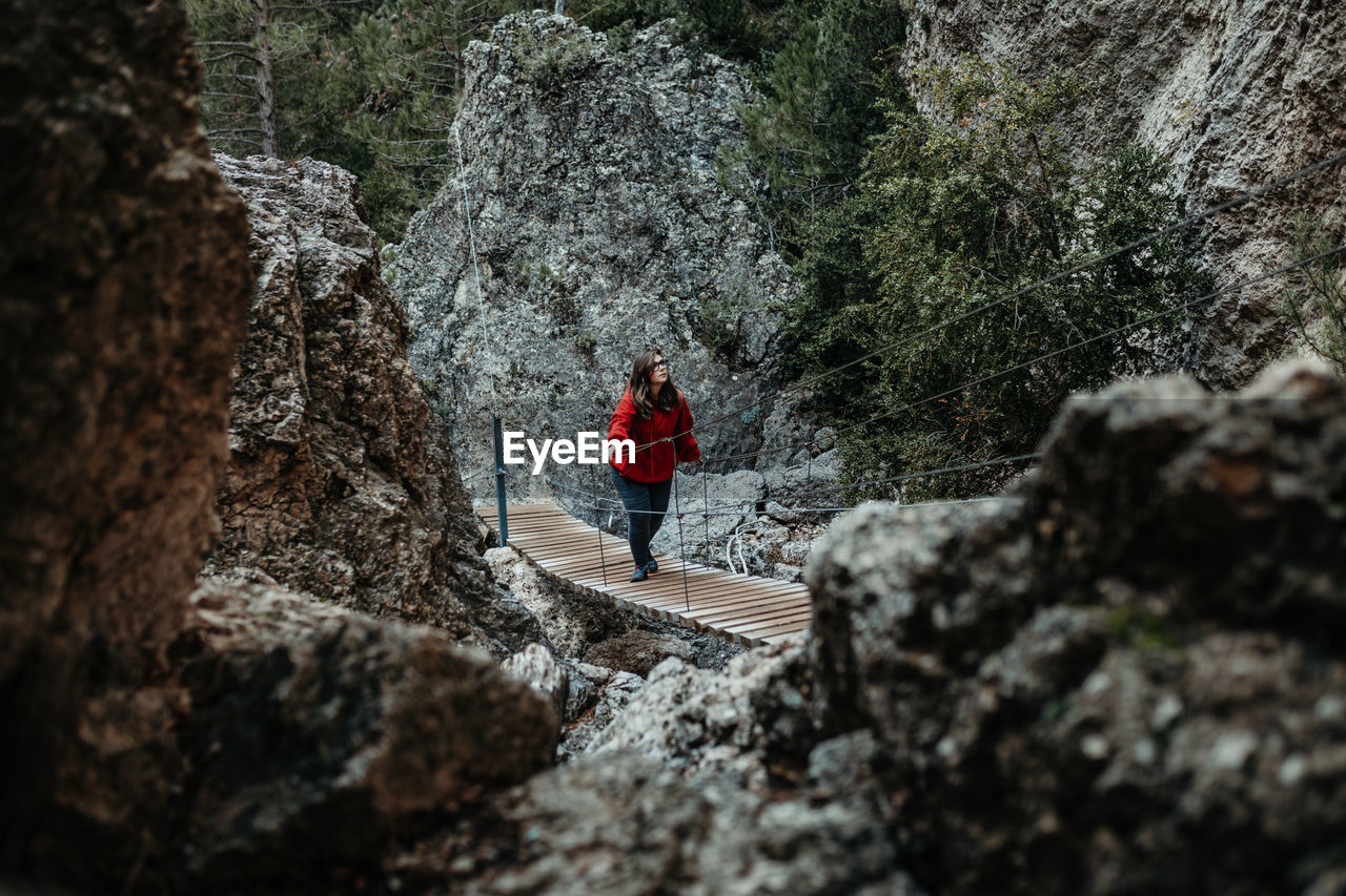 Back view of young tourist with backpack walking on walkway above water near stone wall between forest