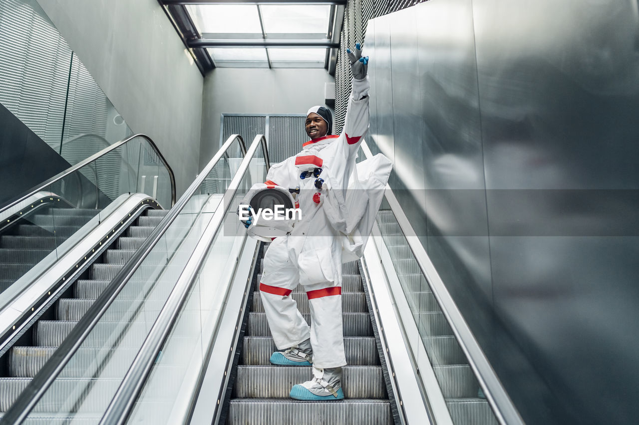 Cheerful astronaut gesturing peace sign while climbing on escalator