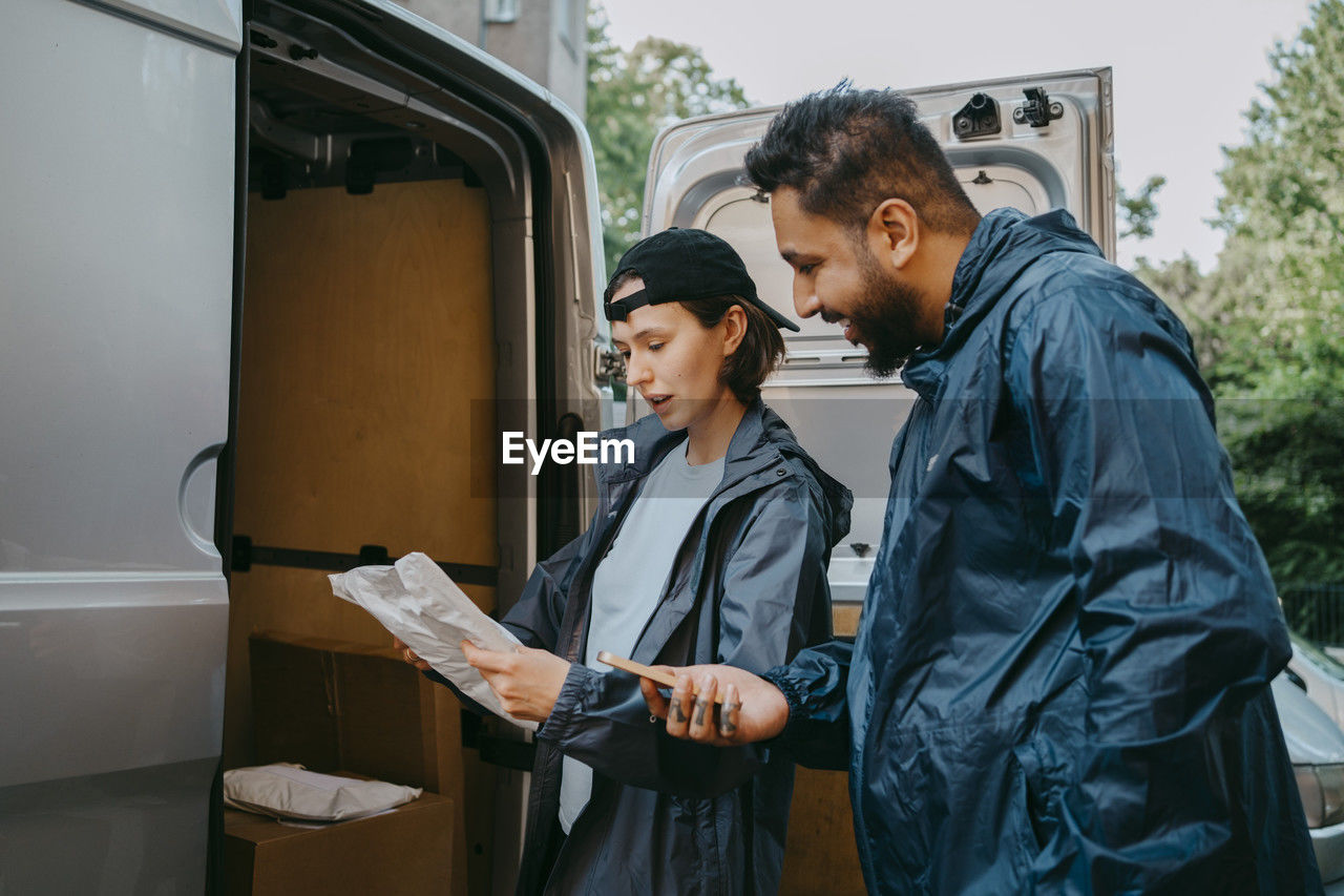 Male and female delivery coworkers reading details on package while standing near van