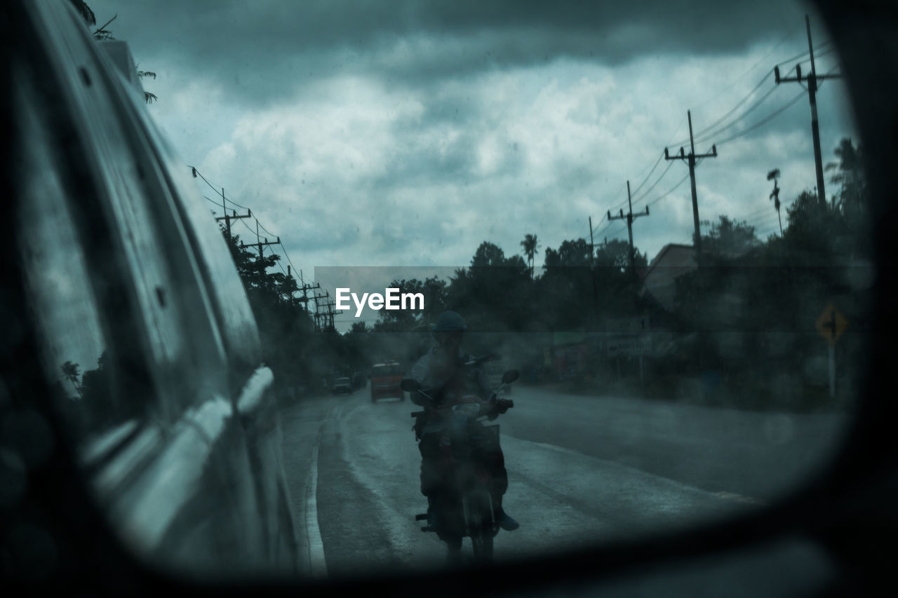 Reflection of man driving scooter on car side-view mirror at road