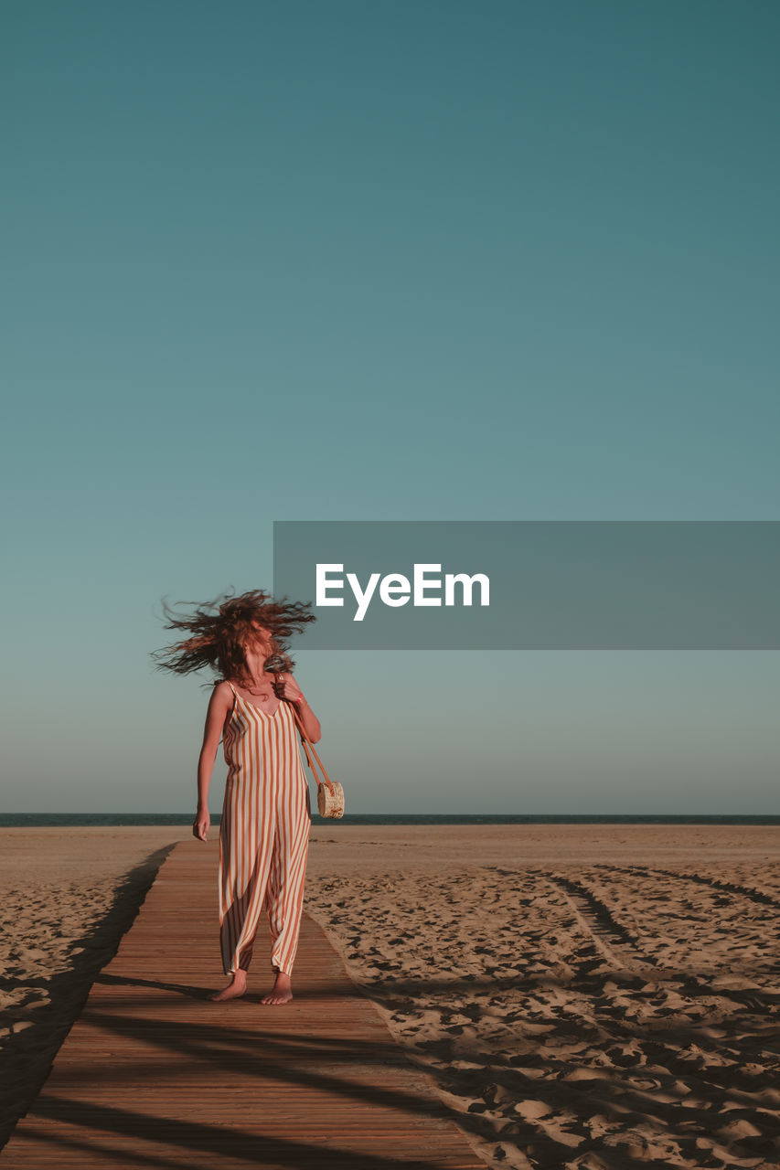 Woman with tousled hair walking on boardwalk at beach against blue sky