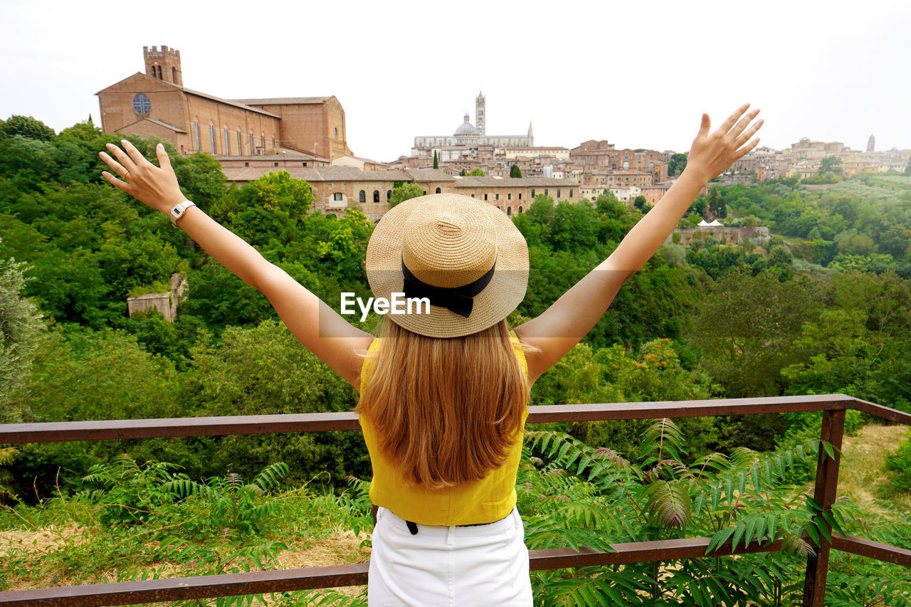 Happy young woman with arms raised on scenic tuscan hills with landmarks in siena, tuscany, italy