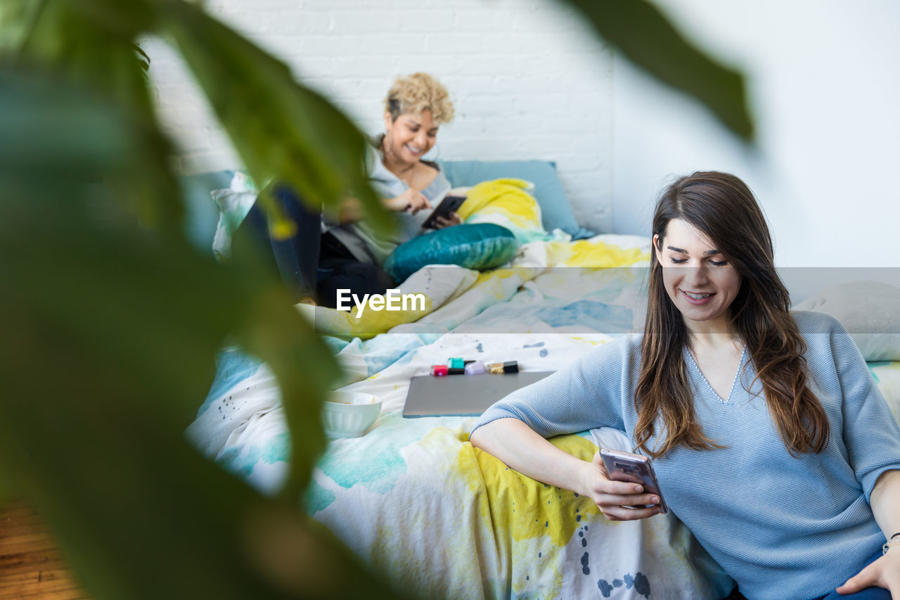 Smiling woman text messaging on smart phone while roommate lying on bed at home