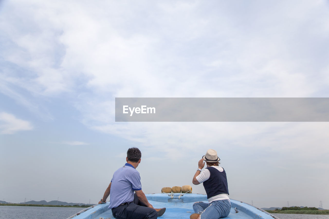 Rear view of men sitting on boat in river against cloudy sky