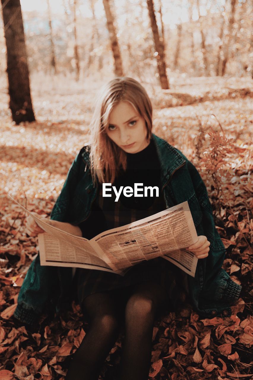 Portrait of young woman holding newspaper while sitting on field during autumn