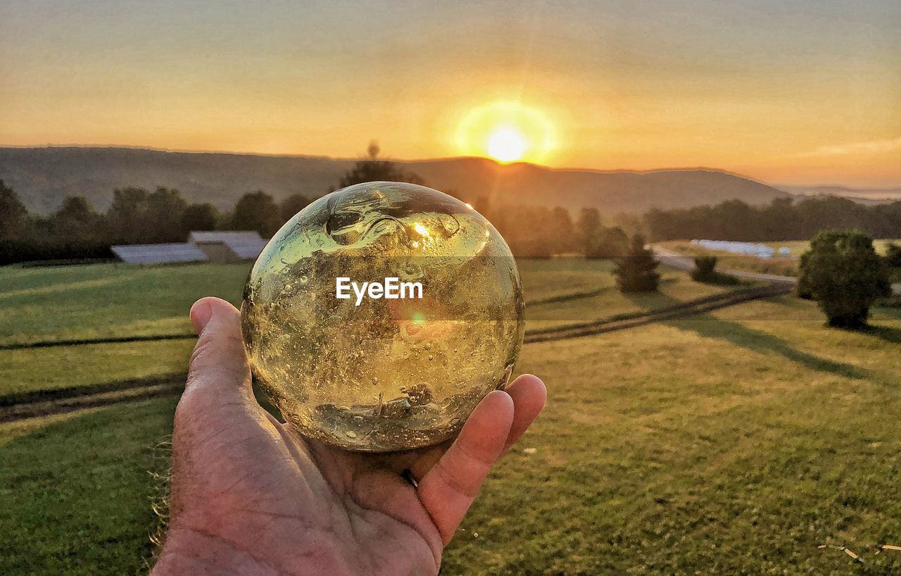 Close-up of hand holding crystal ball against sky during sunset