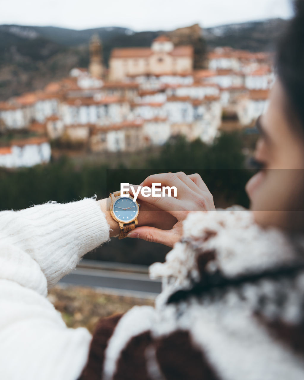 Back view of crop female traveler checking time on trendy wristwatch while standing on hill against old town in cloudy autumn day