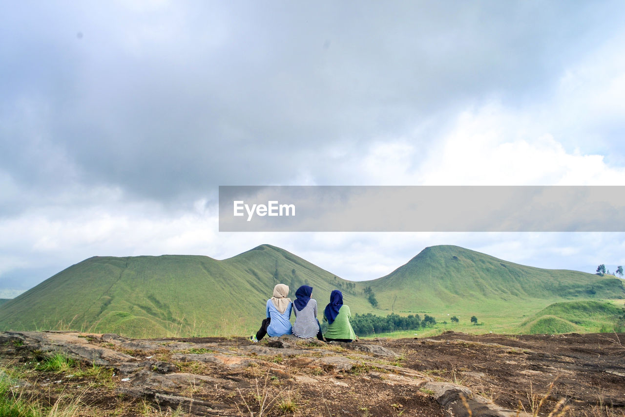 Rear view of three women sitting on mountain against sky