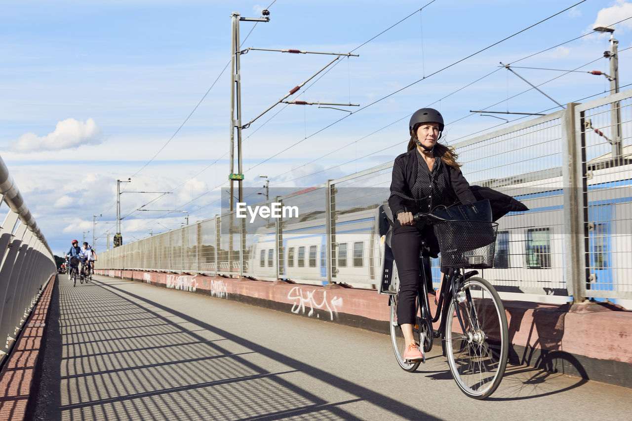 Mature woman cycling on road by fence against sky