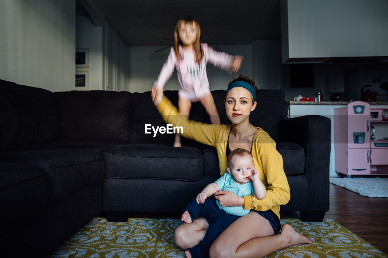 A young mother holding her baby while her toddler jumps on the couch
