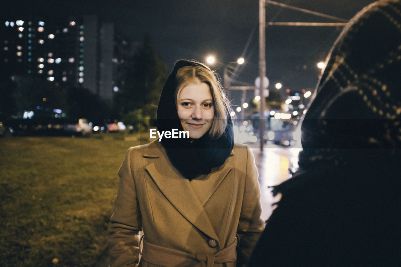 PORTRAIT OF SMILING YOUNG WOMAN STANDING AT NIGHT