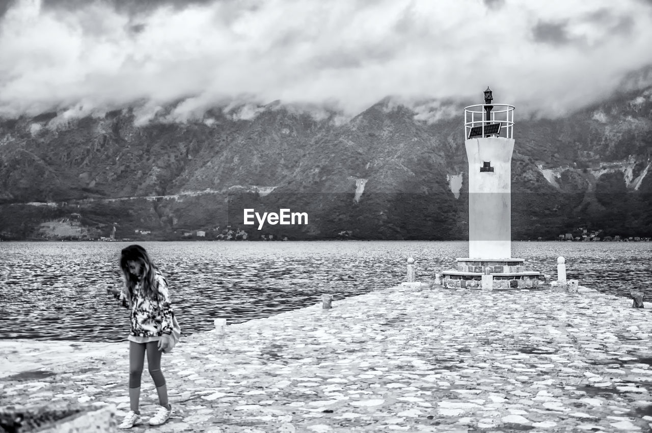 WOMAN STANDING BY LIGHTHOUSE AGAINST SKY ON MOUNTAIN