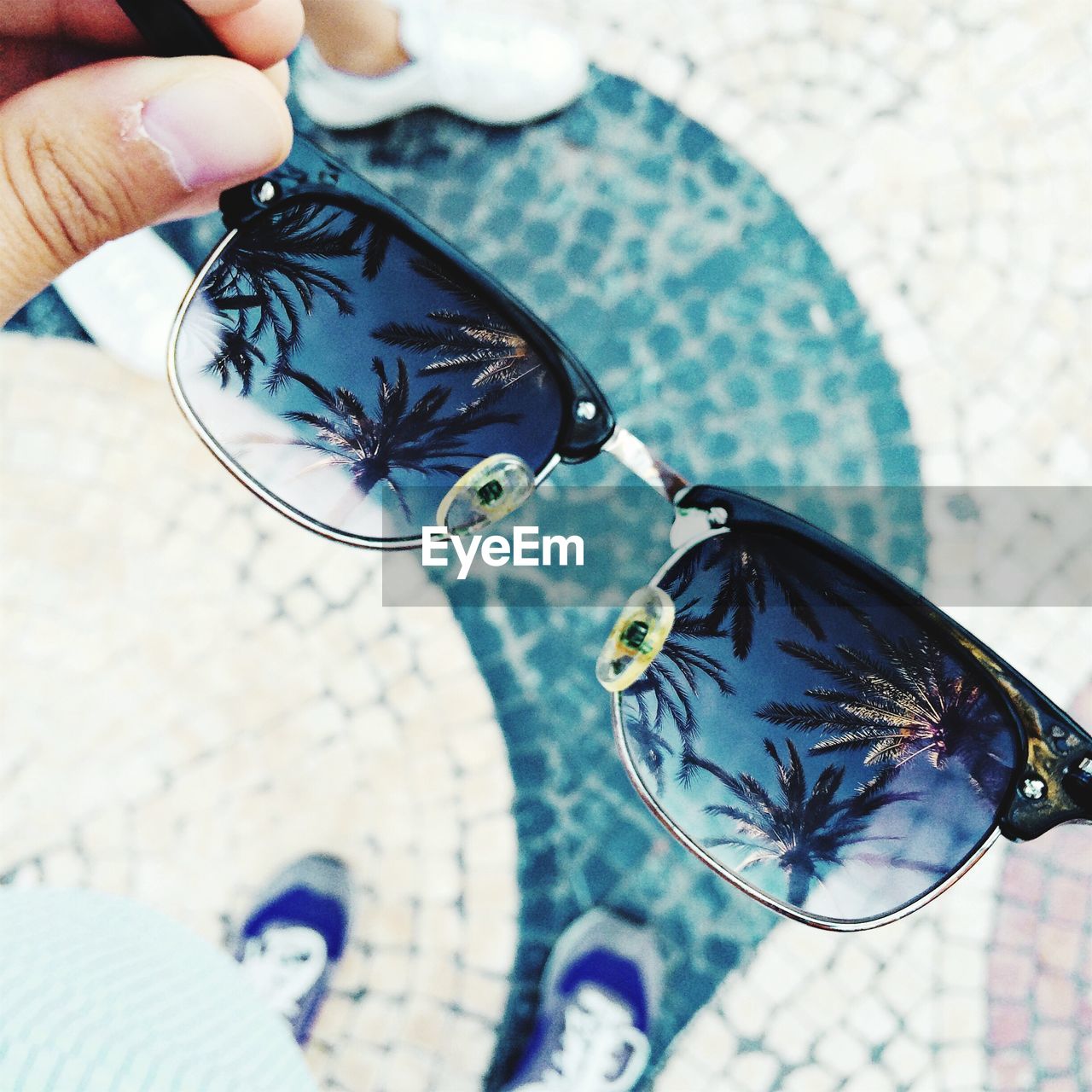 Close-up of hand holding sunglasses with reflection of palm trees