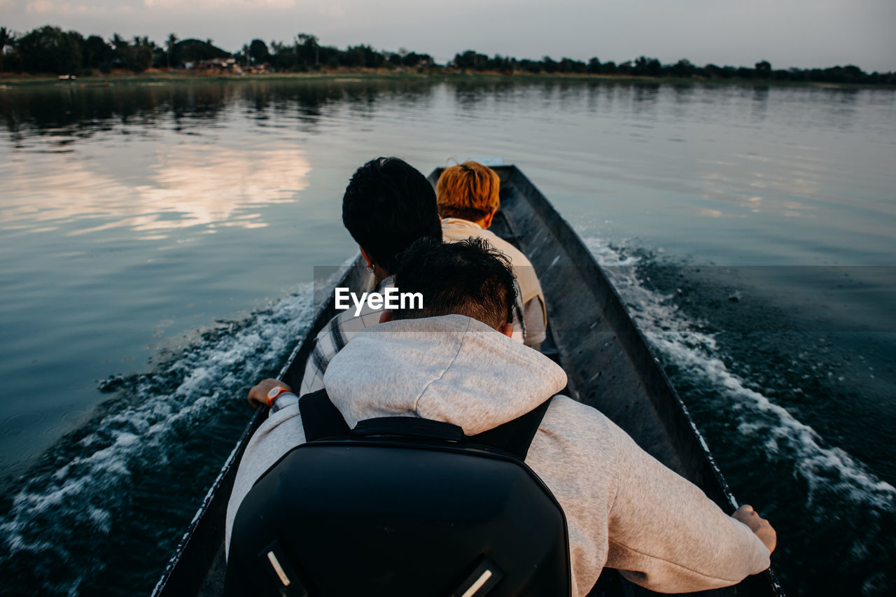 REAR VIEW OF FRIENDS SITTING ON BOAT IN LAKE