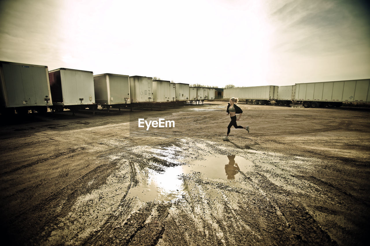 Athlete jogging on muddy field by cargo containers against sky