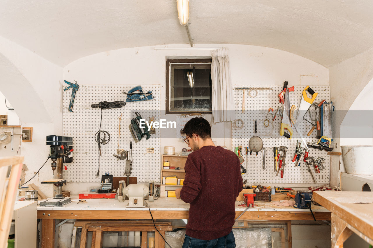 Back view of male woodworker standing at workbench and creating wooden details in bright garage