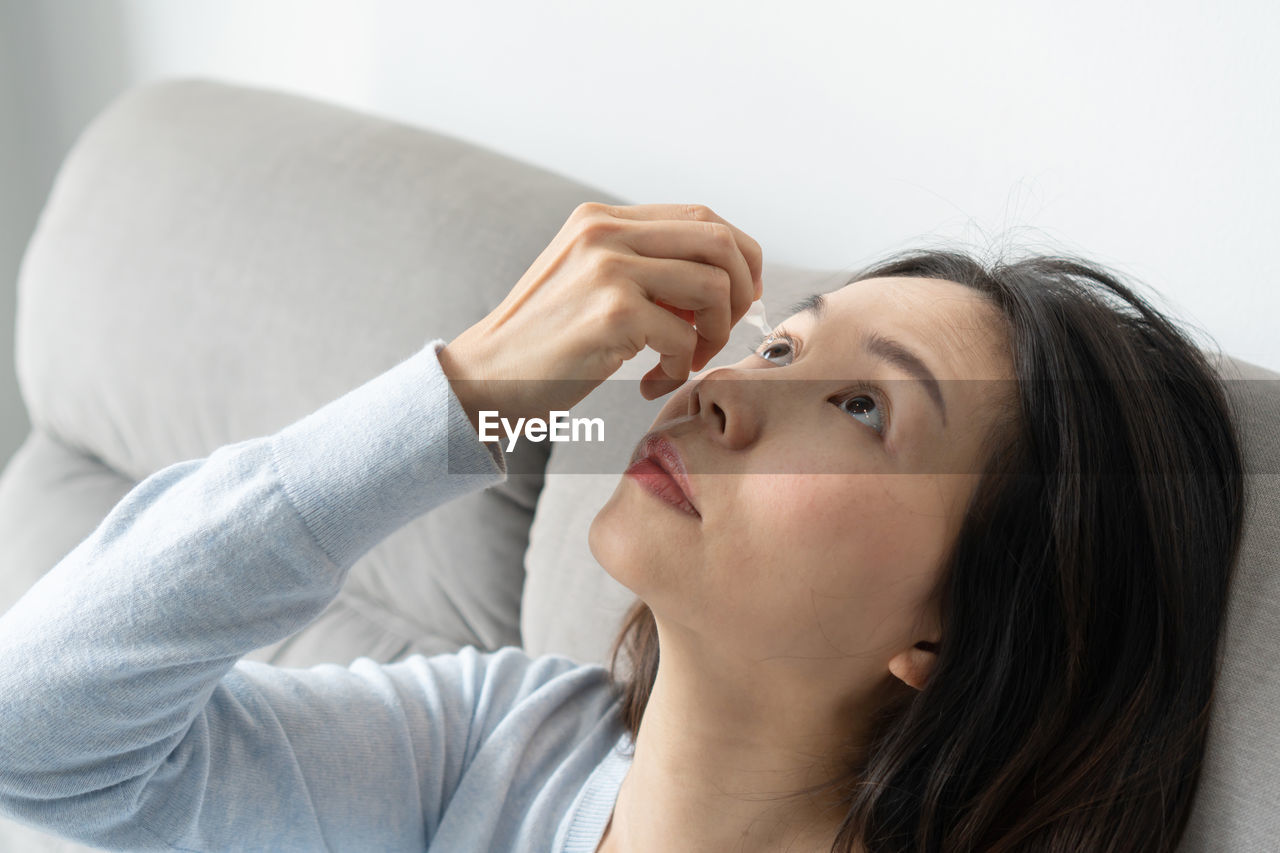 Woman putting eye drop sitting on sofa at home