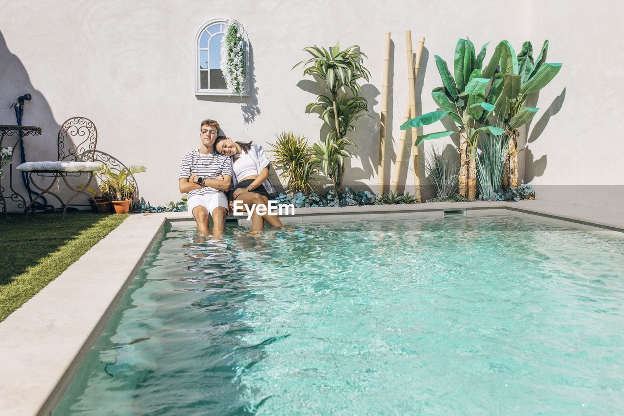 Girl leaning on brother's shoulder by swimming pool in front of wall