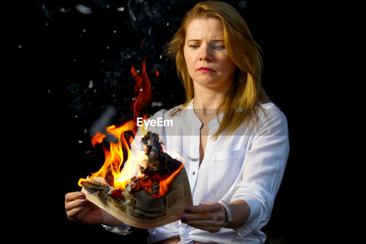 Young woman holding burning newspaper against black background