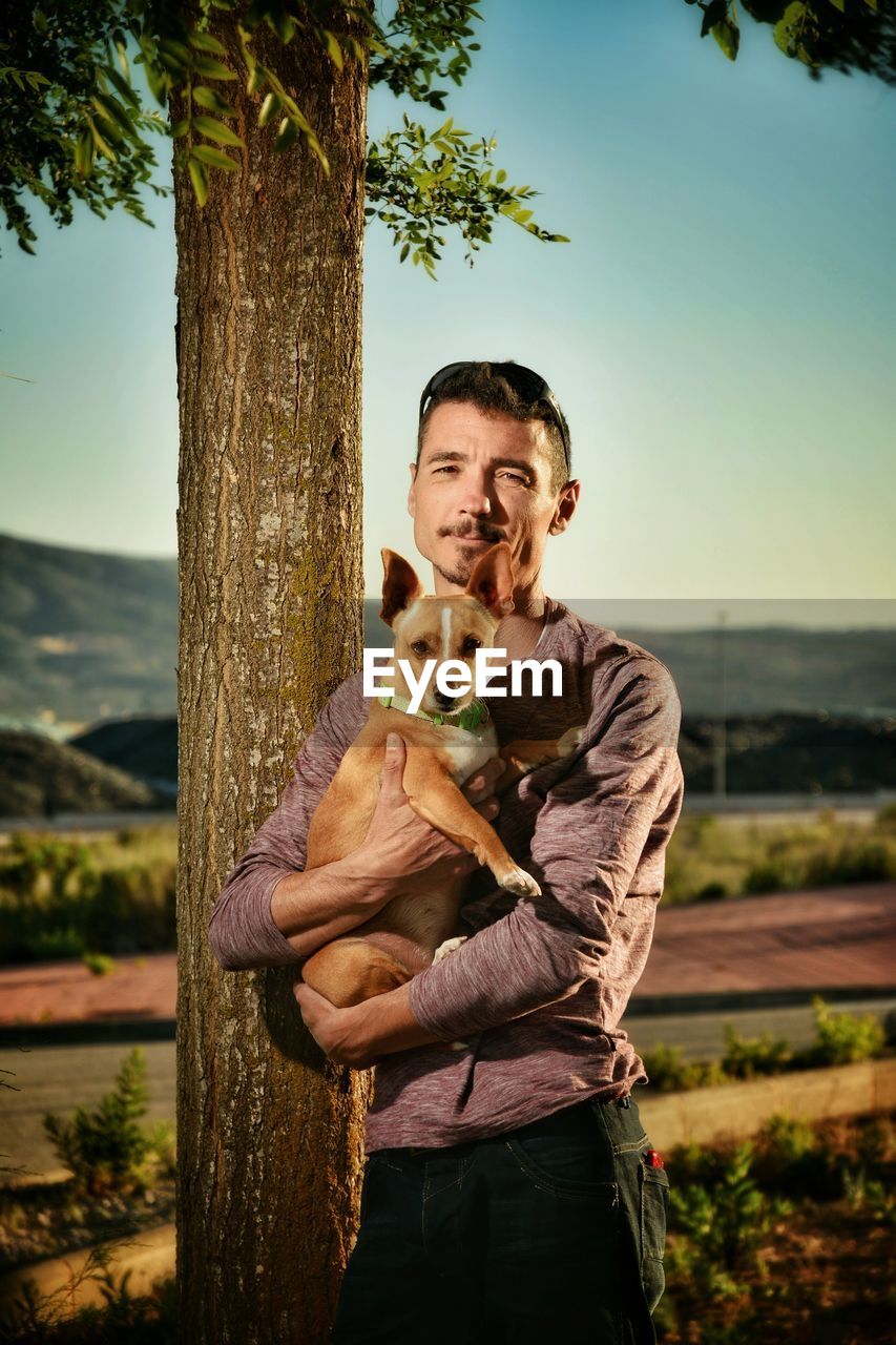 Portrait of mature man holding dog while standing against tree trunk