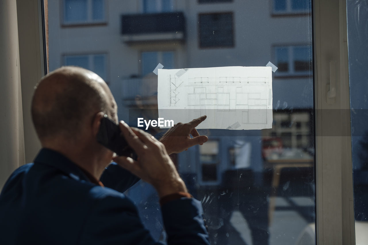 Architect talking through mobile phone analyzing blueprint on window