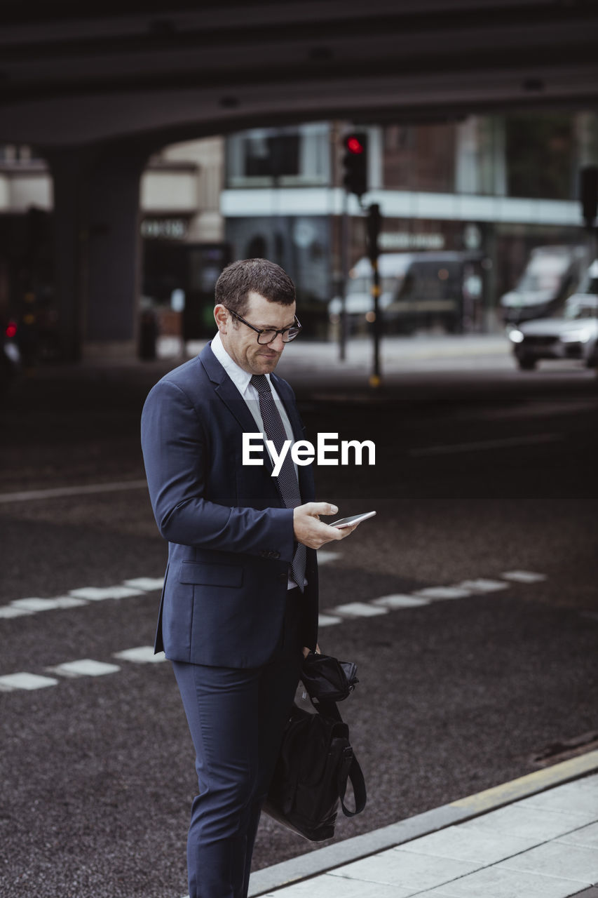 Businessman with bag using smart phone while standing in city