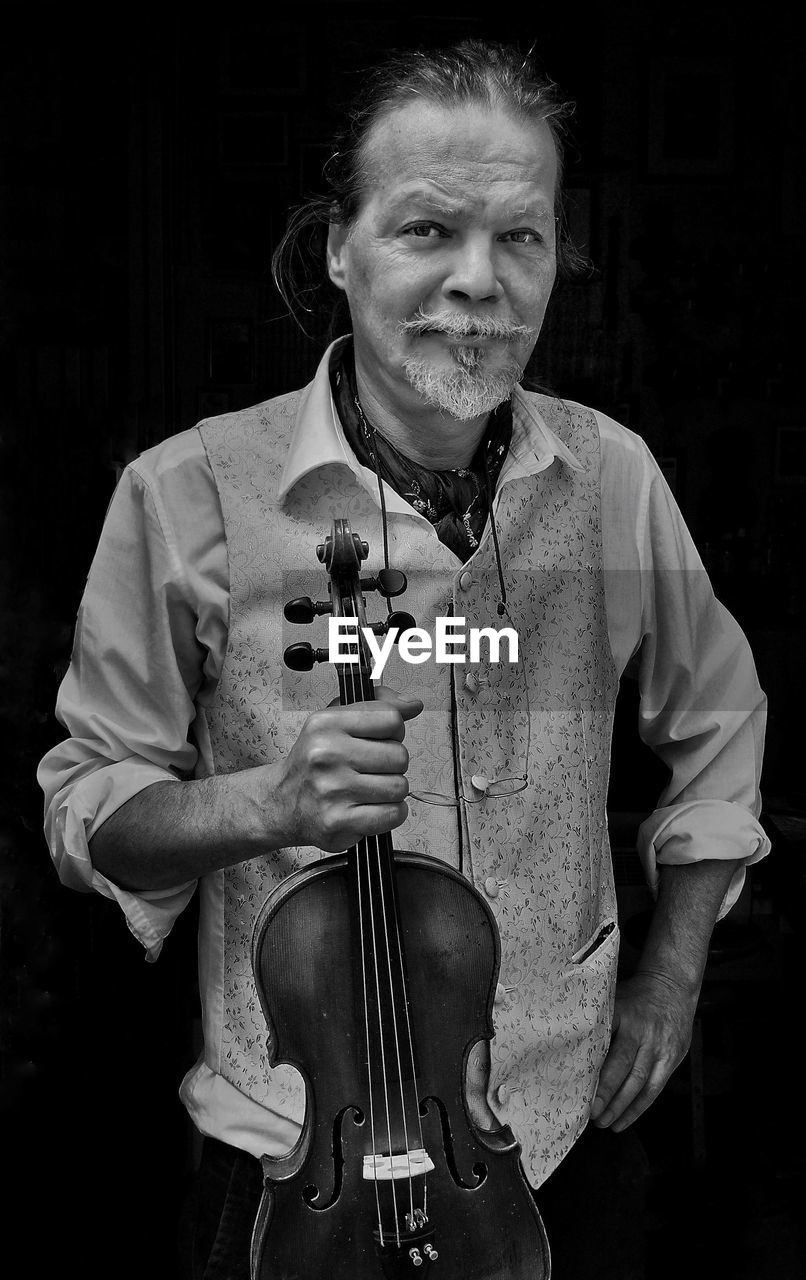 Portrait of man holding violin while standing against black background