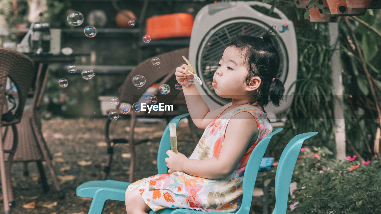 Full length of cute girl blowing bubbles while sitting on chair at yard