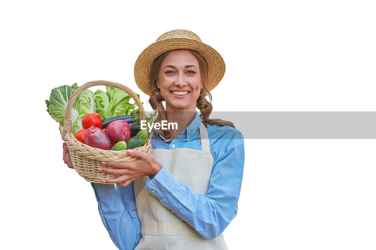 PORTRAIT OF SMILING YOUNG WOMAN HOLDING BASKET AGAINST WHITE BACKGROUND