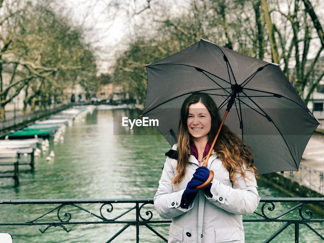 Portrait of mid adult woman holding umbrella while standing on footbridge over canal in city during rainy season