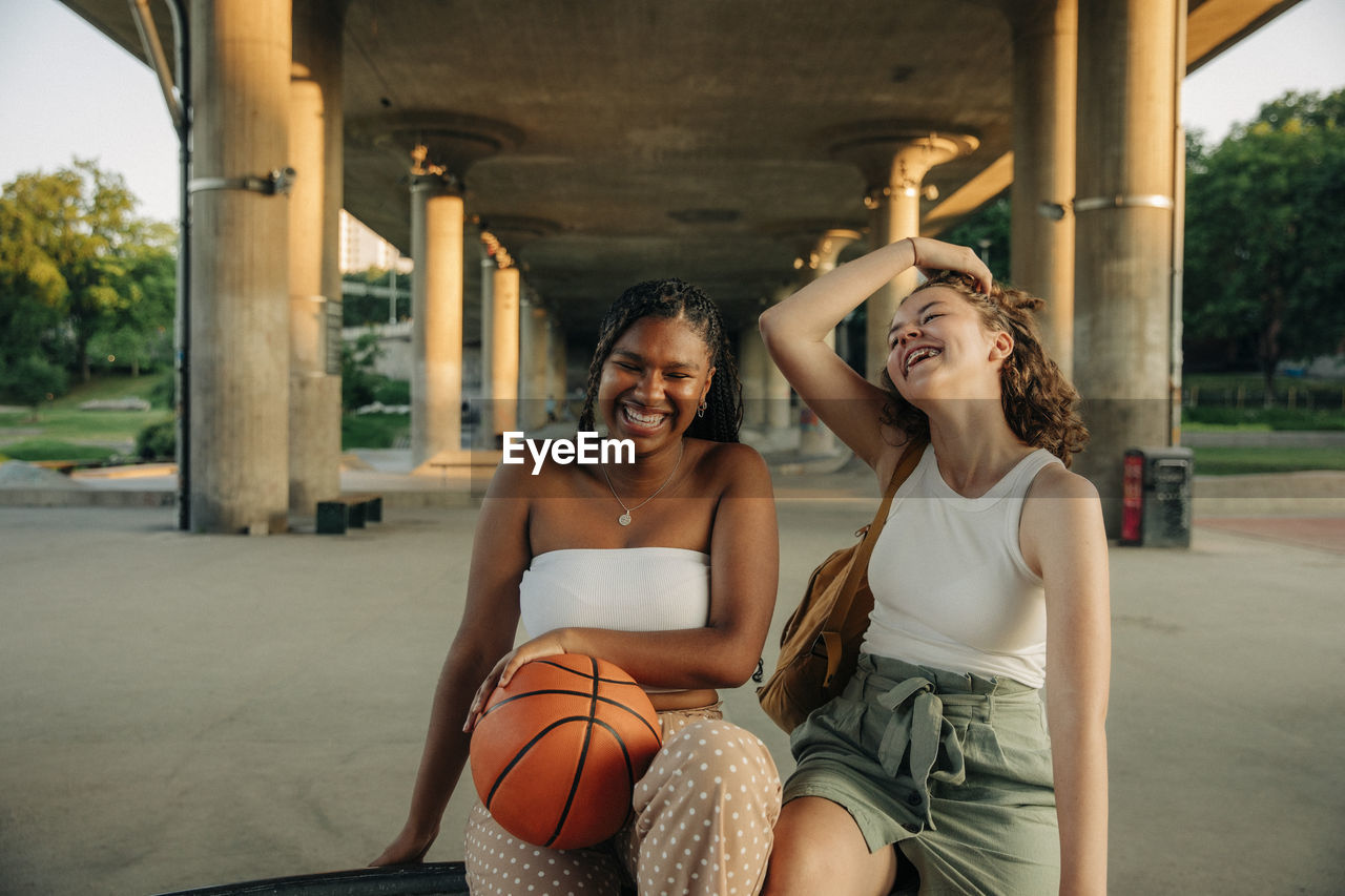 Cheerful teenage girl with basketball sitting by female friend under bridge