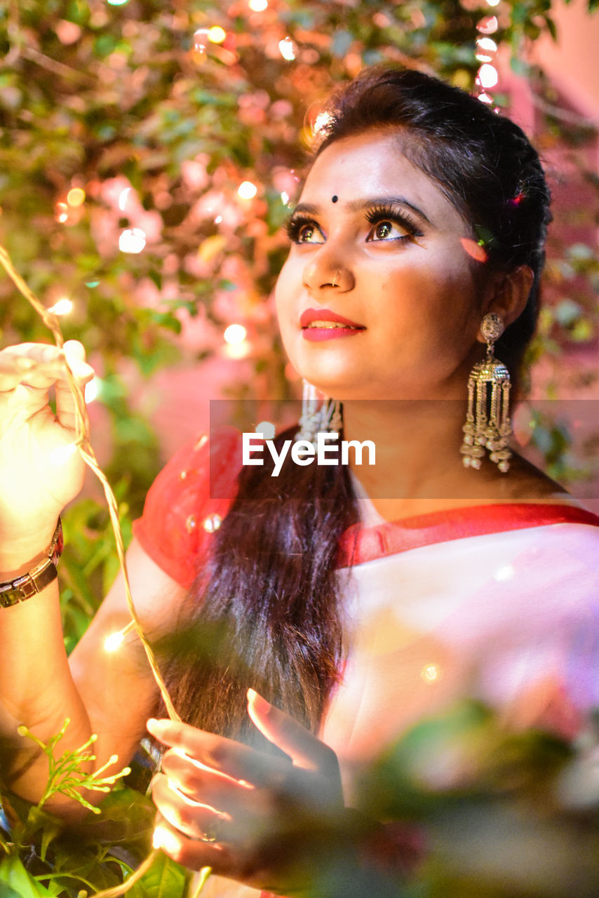 Thoughtful woman holding illuminated string light against plants at night