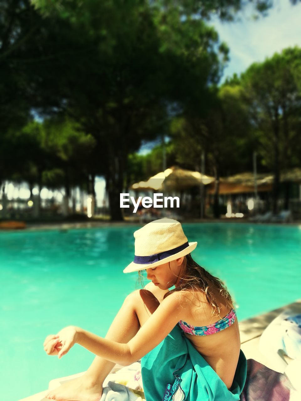 Side view of girl wearing hat while sitting at poolside against trees