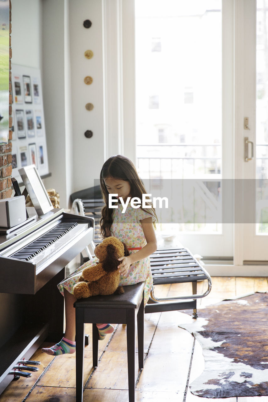 Girl sitting with teddy bear by piano at home