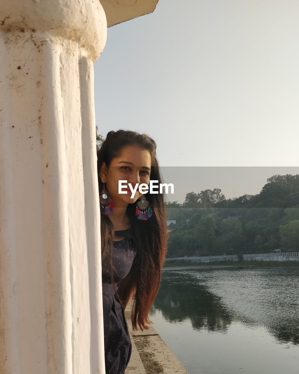 Portrait of smiling young woman standing by lake against sky