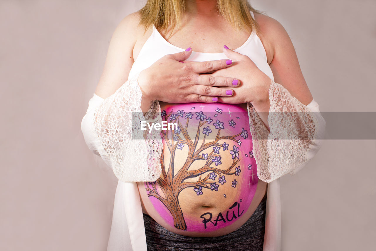 women, adult, pregnant, one person, pink, tattoo, indoors, clothing, female, midsection, studio shot, arm, limb, person, hand, standing, young adult, parent, lifestyles, anticipation, photo shoot, holding, front view, human leg, child, undergarment, skin, blond hair