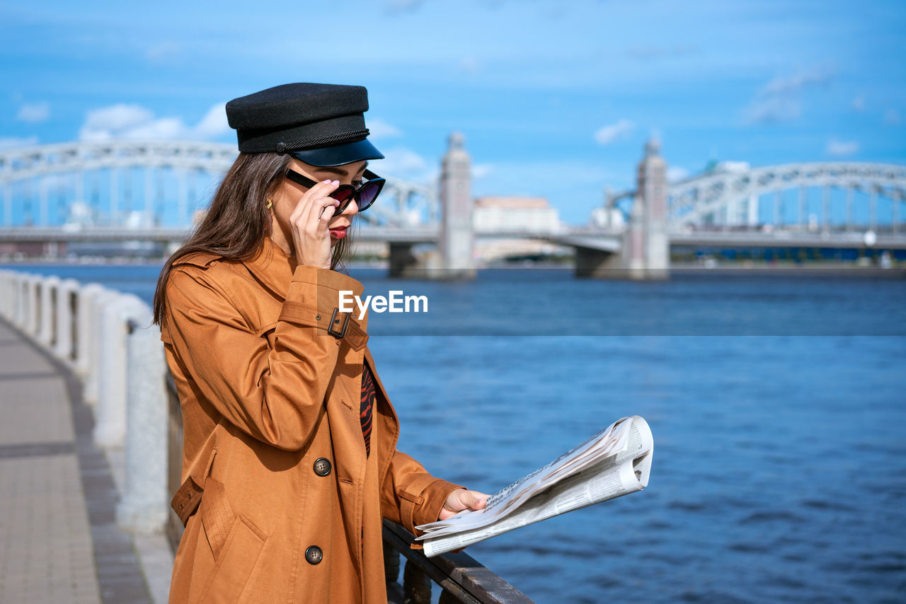 Stylish young woman reading fresh newspaper on embankment of river on sunny day