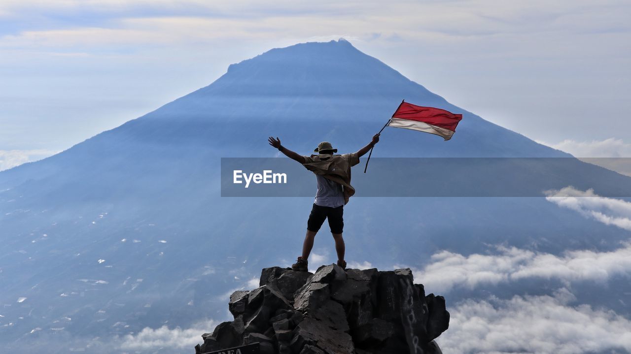 Rear view of man holding flag while standing on rock against sky