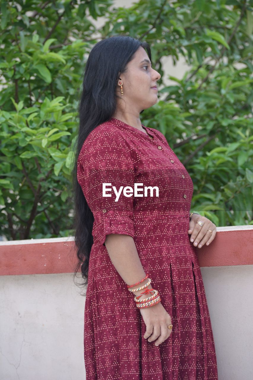 one person, pink, women, adult, maroon, dress, pattern, standing, photo shoot, clothing, three quarter length, young adult, looking, lifestyles, hairstyle, red, looking away, fashion, magenta, textile, plant, day, nature, female, gown, outdoors, waist up, black hair, outerwear, long hair, portrait, casual clothing, purple, contemplation, leisure activity, brown hair, person, smiling, emotion, side view