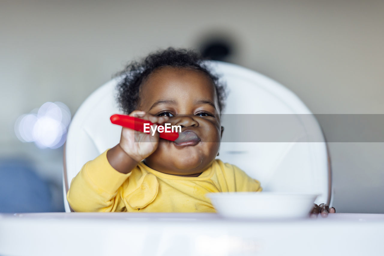 Baby girl eating with spoon while sitting on high chair at home