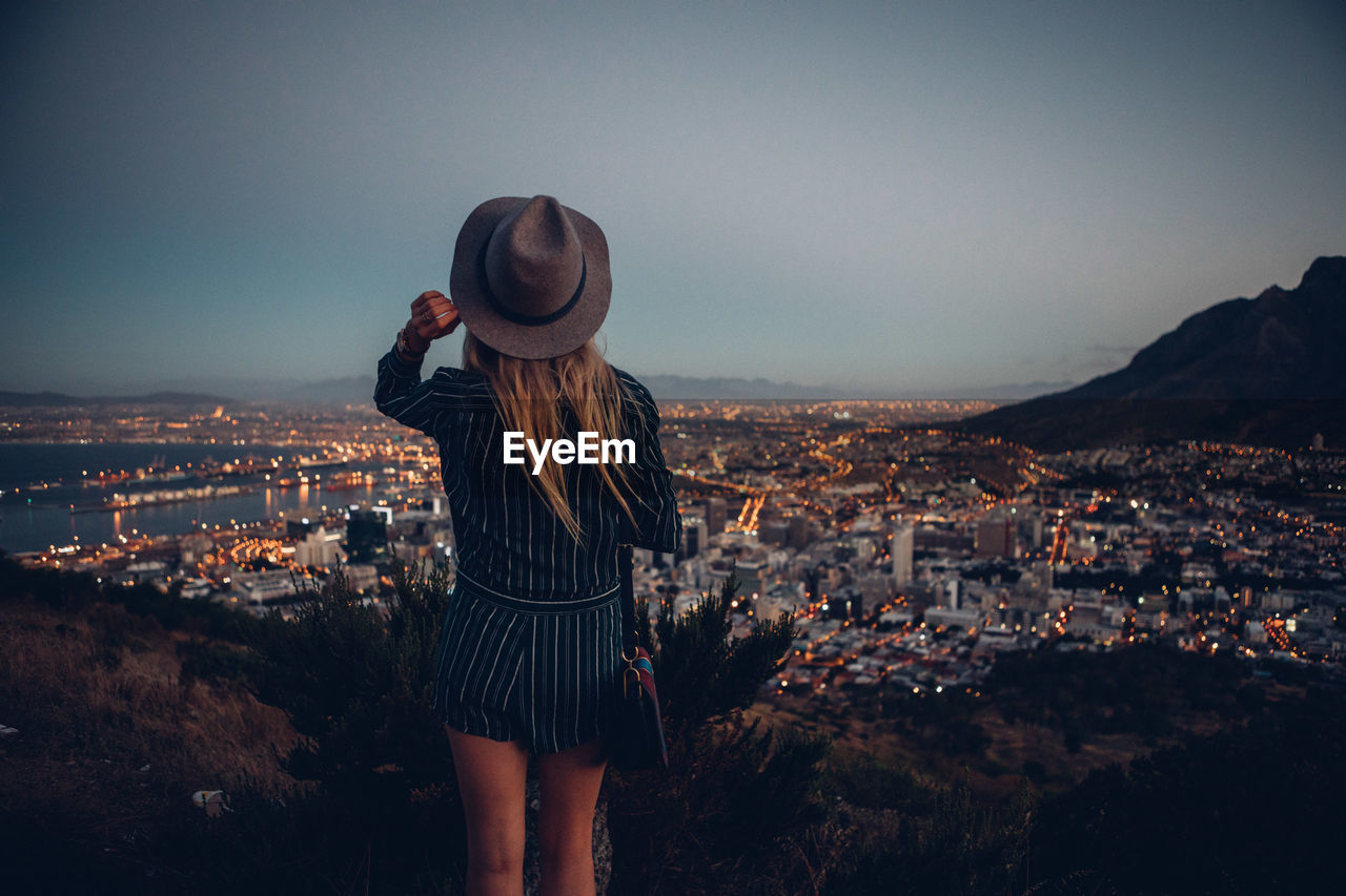 Rear view of woman by illuminated cityscape against clear sky during dusk