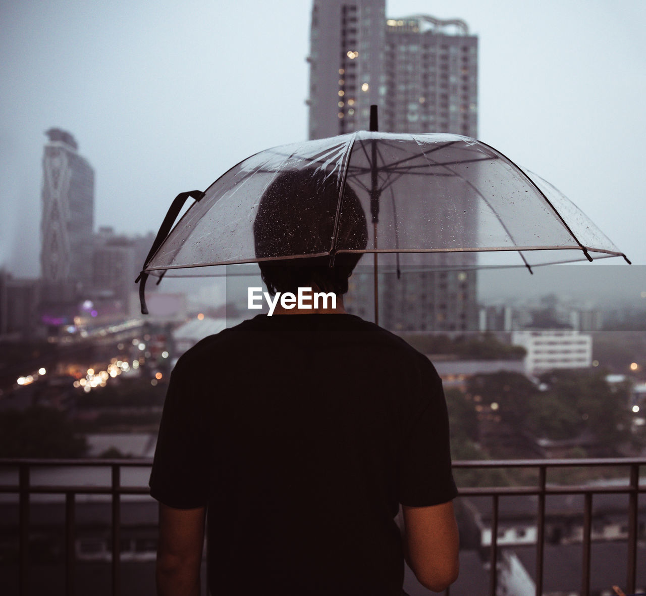 REAR VIEW OF MAN STANDING AGAINST CITYSCAPE DURING RAINY DAY