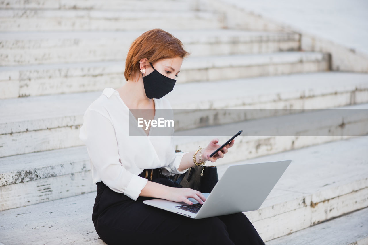 Concentrated businesswoman in elegant clothes and medical mask sitting on stairs on street and working on remote project while using laptop and smartphone